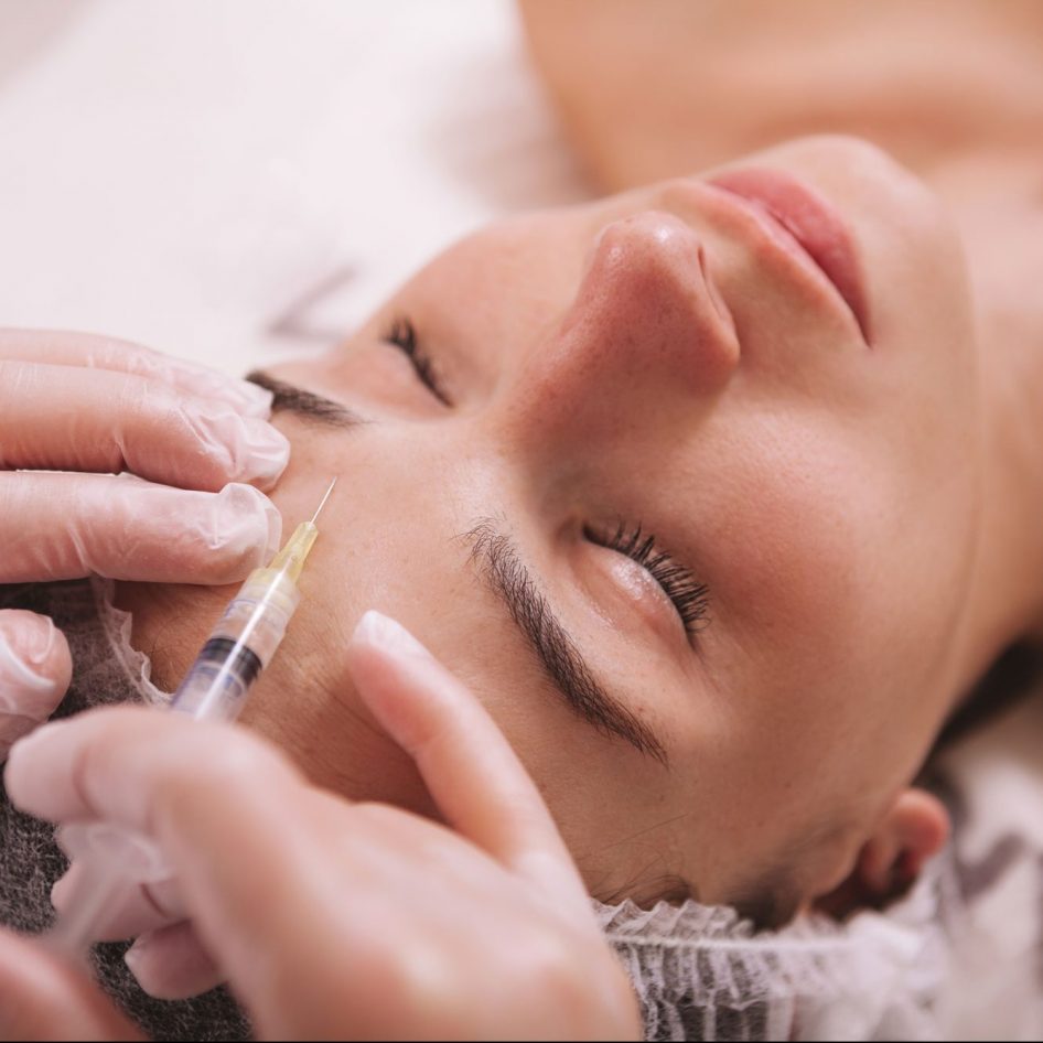 Top view close up of a woman receiving filler injections in forehead. Professional cosmetologist injecting hyaluronic acid into skin of female client